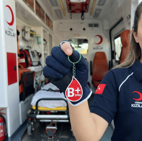 Ambulansta NFC Kan Grubu Anahtarlığı Kullanımı Ambulansın içinde tıbbi ekipmanların ve sedyenin önünde duran Türk Kızılay üniformalı bir sağlık görevlisi, elinde B+ pozitif kan gruplu kırmızı NFC akıllı anahtarlığı tutarak gösteriyor.